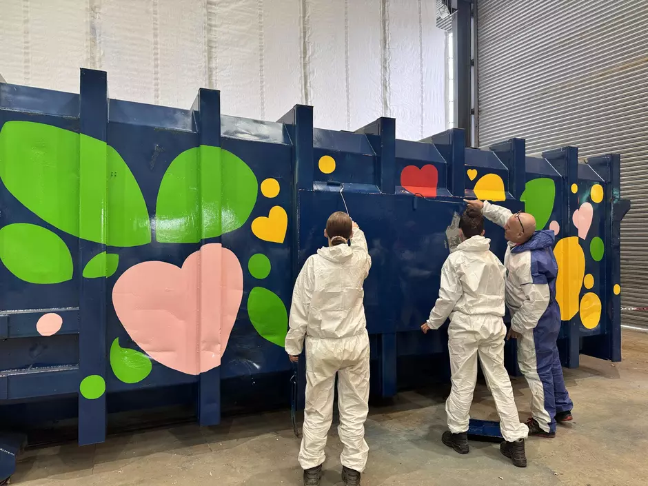 3 people in white coveralls painting a blue container with brightly coloured patterns. 