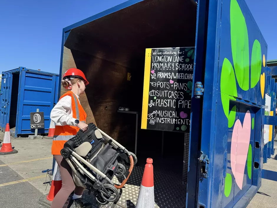 Member of staff carrying a pram into a brightly painted GoodBox container with a chalkboard listing items collected for reuse